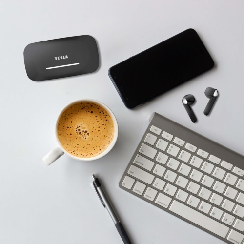 Pracovní prostor s klávesnicí, šálkem kávy a mobilním telefonem, doplněný o bezdrátová Bluetooth sluchátka TESLA Sound EB20 Black Raven a jejich nabíjecí pouzdro.